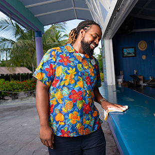 black male bartender, wearing a Hawaiian shirt and smiling in front of a resort bar