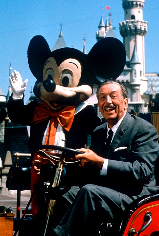Walt Disney riding in a cart with a person wearing a Mickey Mouse costume, at a Disney park