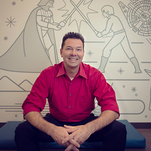 Man with brown hair wearing a red collared shirt smiling while sitting in front of a wall with outlined scenes from Star Wars