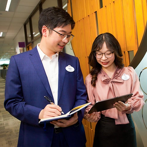 Young Asian man and woman in business attire standing in a corridor, the man taking notes while looking at the woman's tablet screen