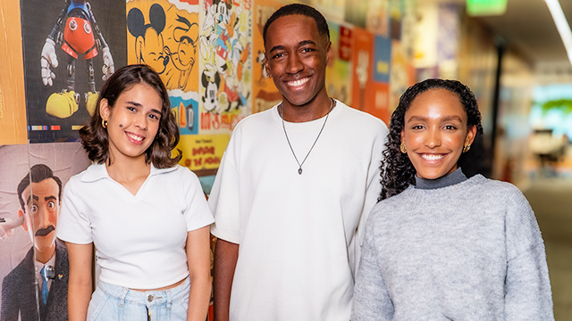 Three TWDC Latin America interns smiling in front of a wall with various Disney movie and show posters