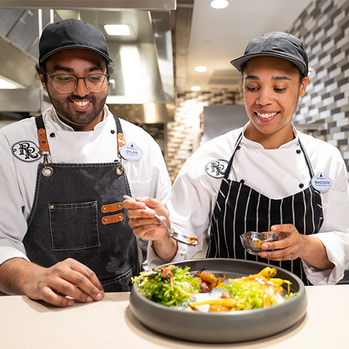 Two culinary staff members in a kitchen smiling while taste testing a salad