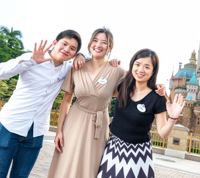 Three interns smiling and waving in front of a Cinderella's Castle in Hong Kong Disneyland Resort