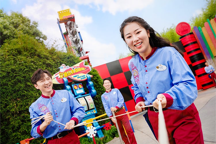 three young Disney cast members in uniform/costume, smiling and holding a rope at a Disney park attraction