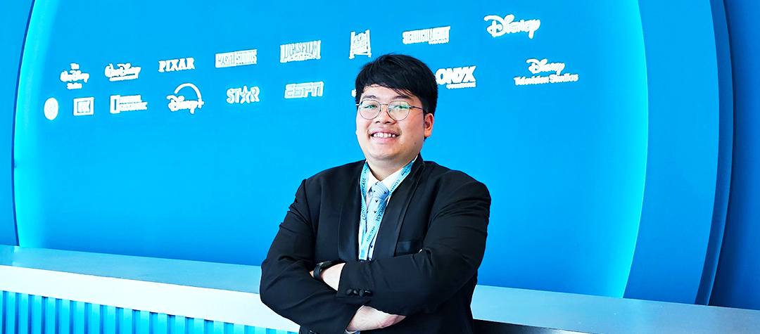 Singapore male intern wearing glasses, a collared shirt and black blazer smiling in front of a wall of The Walt Disney Company business logos