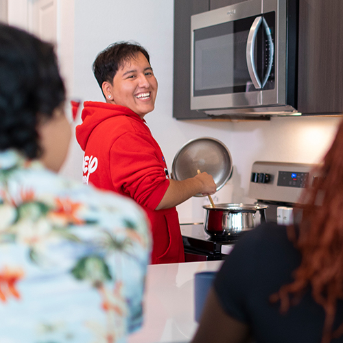 Young hispanic male wearing a Disney sweatshirt, cooking for family at home and smiling