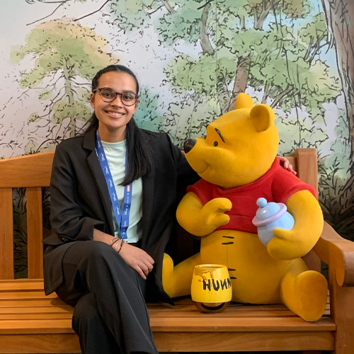 Disney UK Intern Bibechana smiling with long black hair, glasses and a brown sweater. Chip, Dale and Pluto animated characters are in her background.