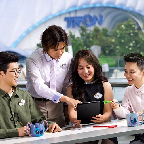 group of young employees in business attire sitting at a table in a Disney park, looking at a tablet