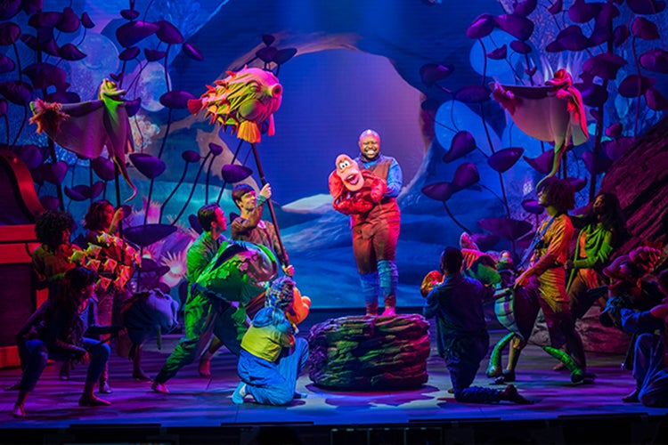 On stage production of the Little Mermaid with cast, colorful fish and Sebastian the crab