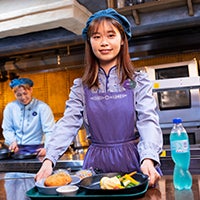 Female Hong Kong Disneyland Food & Beverage Services Host  wearing a purple dress and smiling while holding a tray of food