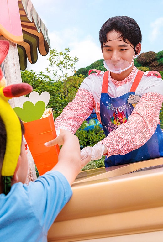 Young male Disney park employee at a popcorn stand, handing popcorn to a child