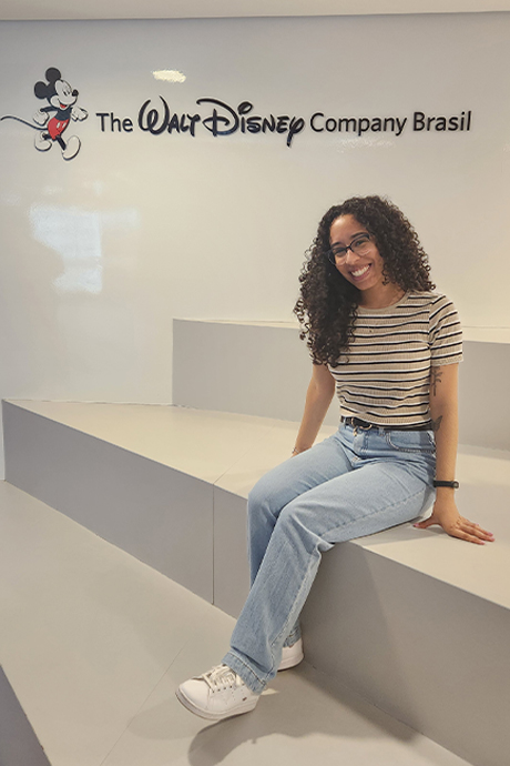 Brasil Apprentice Vitoria wearing a striped shirt and jeans smiling in front of a Walt Disney Company Brasil sign