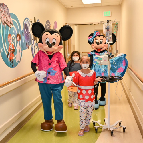 Mickey and Minnie walking with two children wearing masks with a portable IV drip down the hall of a hospital