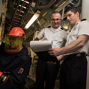 two male engineering officers inspecting paperwork while an engineering crew member performs work on the ship's engine