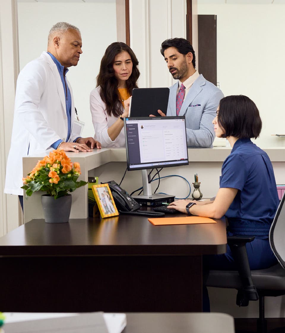 Two FRM's consult with a doctor and a nurse in front of a tablet behind the front desk of a doctor's office.