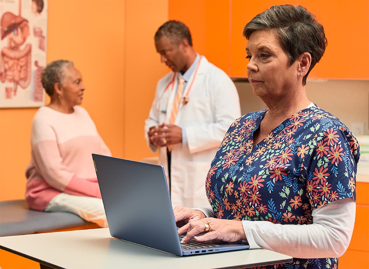 Care team member working at mobile workstation with provider speaking with patient in the background.