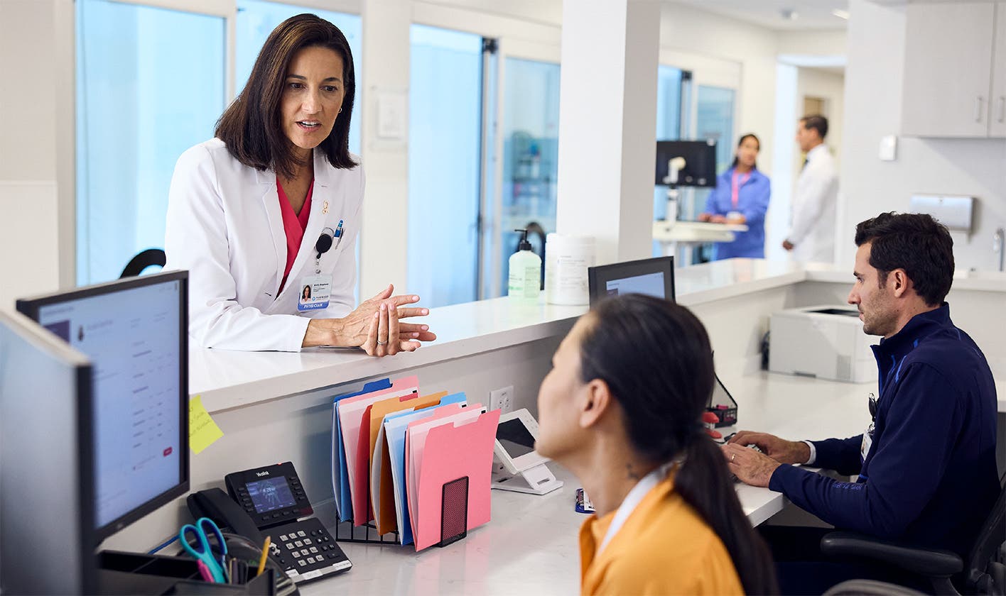 A provider, leaning over the counter, consulting with a care team member at their workstation in a medical clinic.