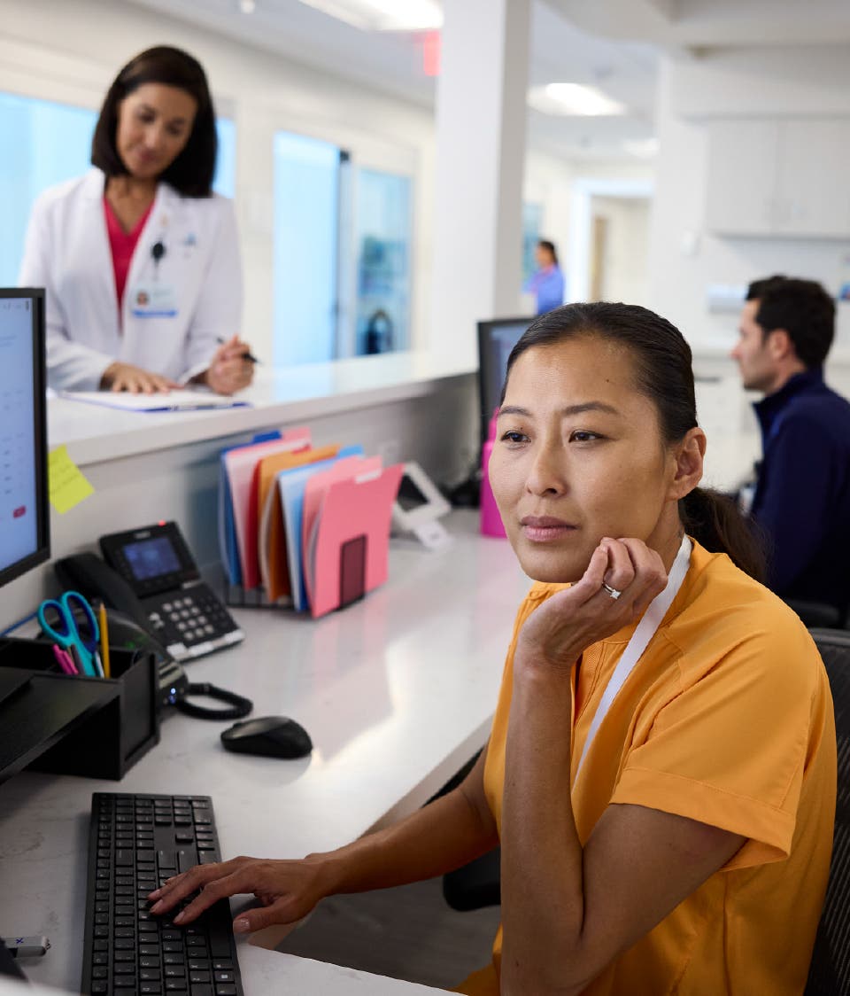 A nurse sitting at a computer, studying a chart while a doctor in a white coat stands behind her reviewing a paper document in a doctor's office