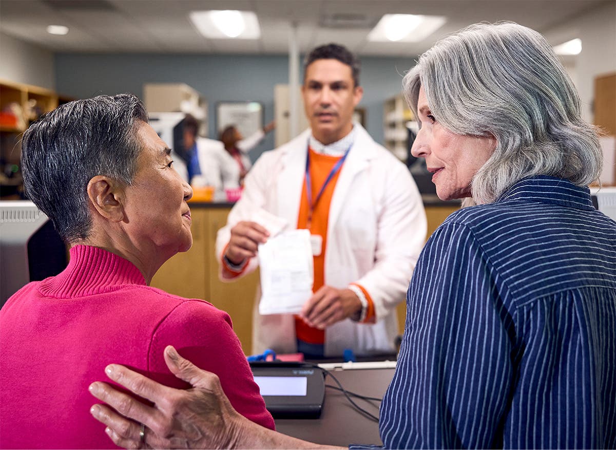 Patients at the pharmacy counter look at each other while pharmacist in background provides information regarding their prescription