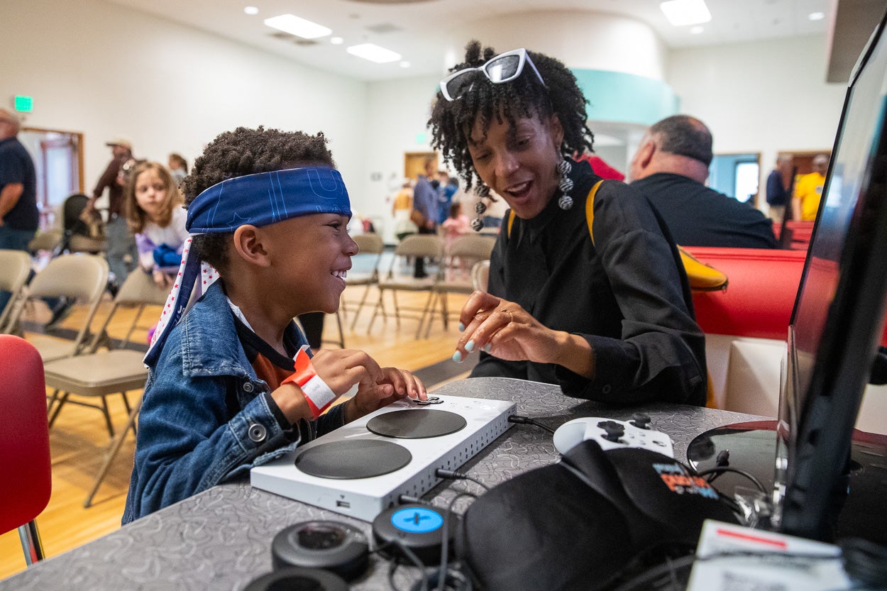 a woman and child sit in front of an adapted video game controller and watch a screen touching the controller.
